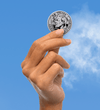 Hand holding a silver coin with '25 Years' and a face on it against a blue sky background