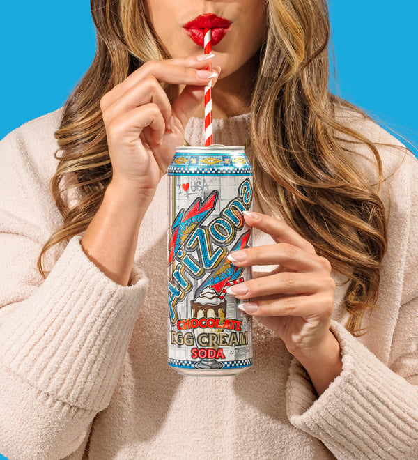Person holding a can of Arizona Egg Cream Soda with a colorful label against a blue background