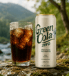 A can of Green Cola Zero soda stands next to a glass of dark cola with ice cubes on a mossy outdoor surface, with a blurred mountain and river background.