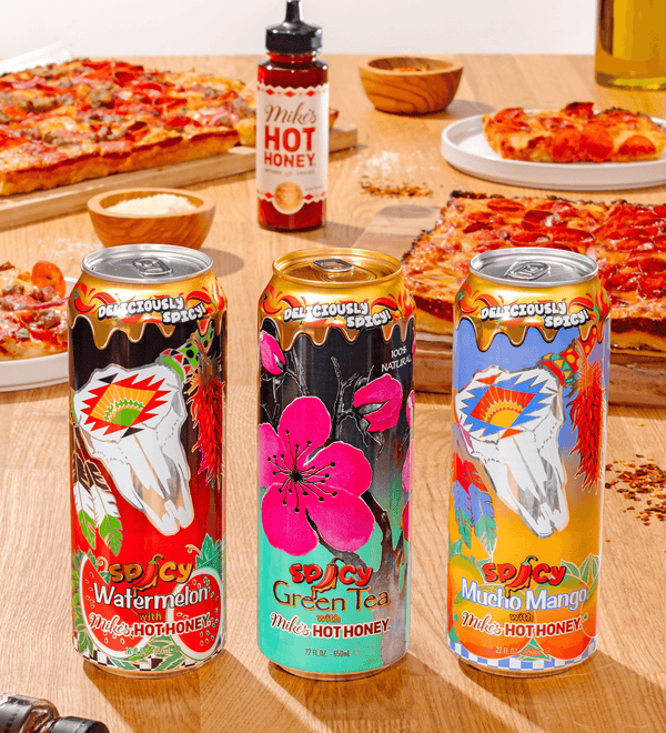 Three cans of spicy AZ with colorful labels on a table with pizzas and a bottle of Mikes hot honey sauce.