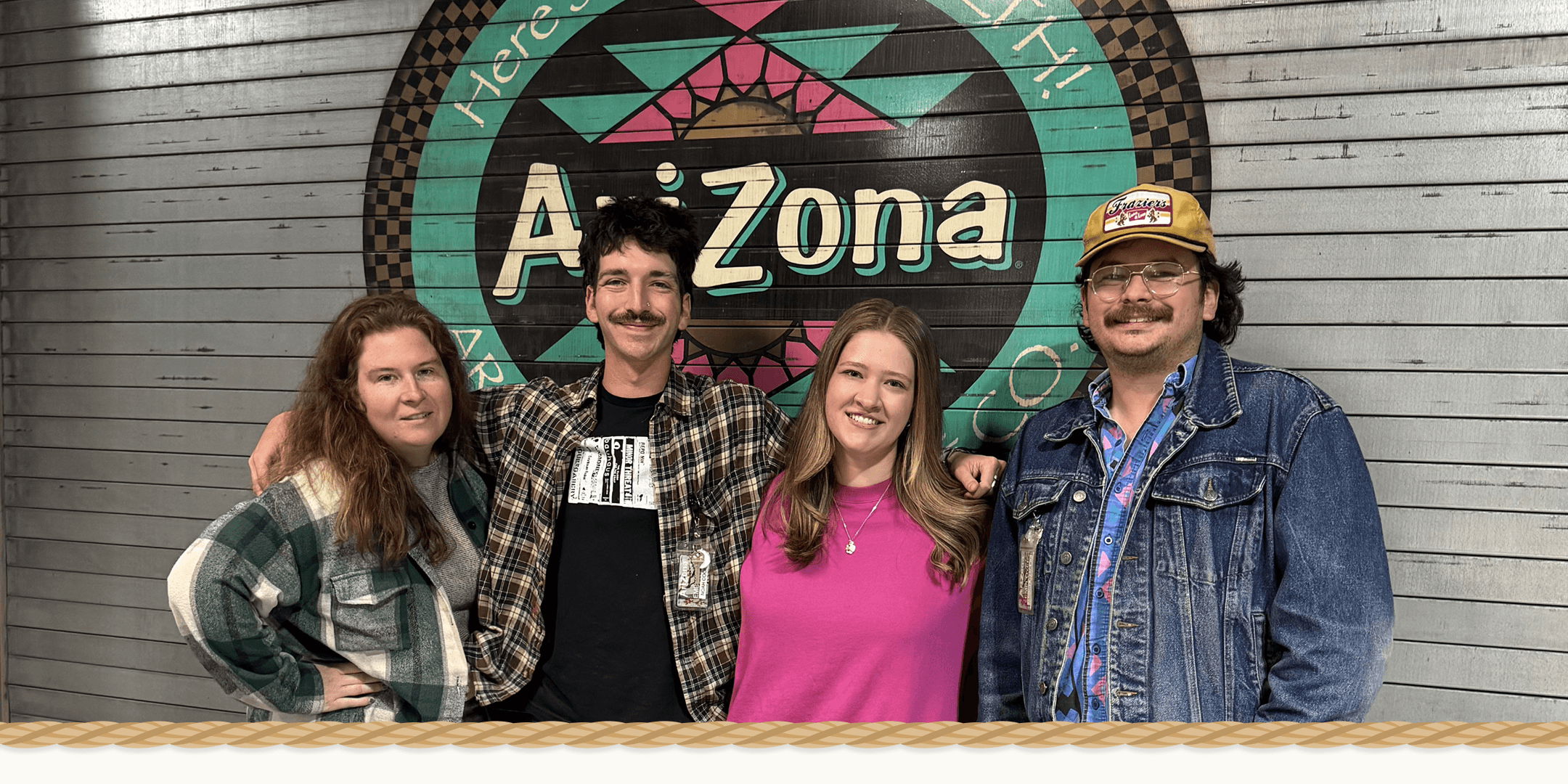 Four people standing together in front of a large circular Arizona-themed sign on a gray wood-paneled wall.