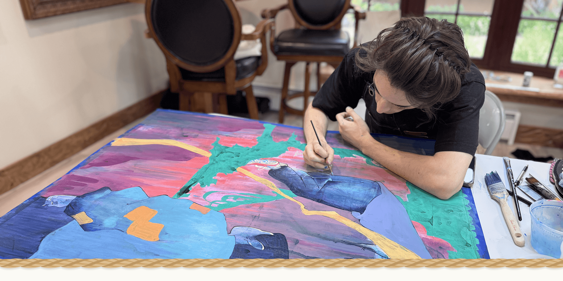 A person painting a large abstract canvas in a studio or home workshop setting. The artwork features bold brushstrokes in blue, green, pink, purple, and yellow.
