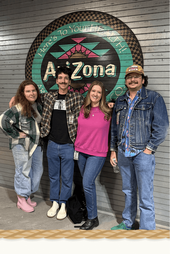 Four people standing in front of a large circular 'Arizona' sign on a gray slatted wall. 