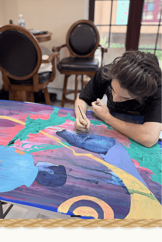 A person painting a colorful abstract artwork on a large canvas table in an indoor studio or gallery space with wooden chairs and windows in the background.