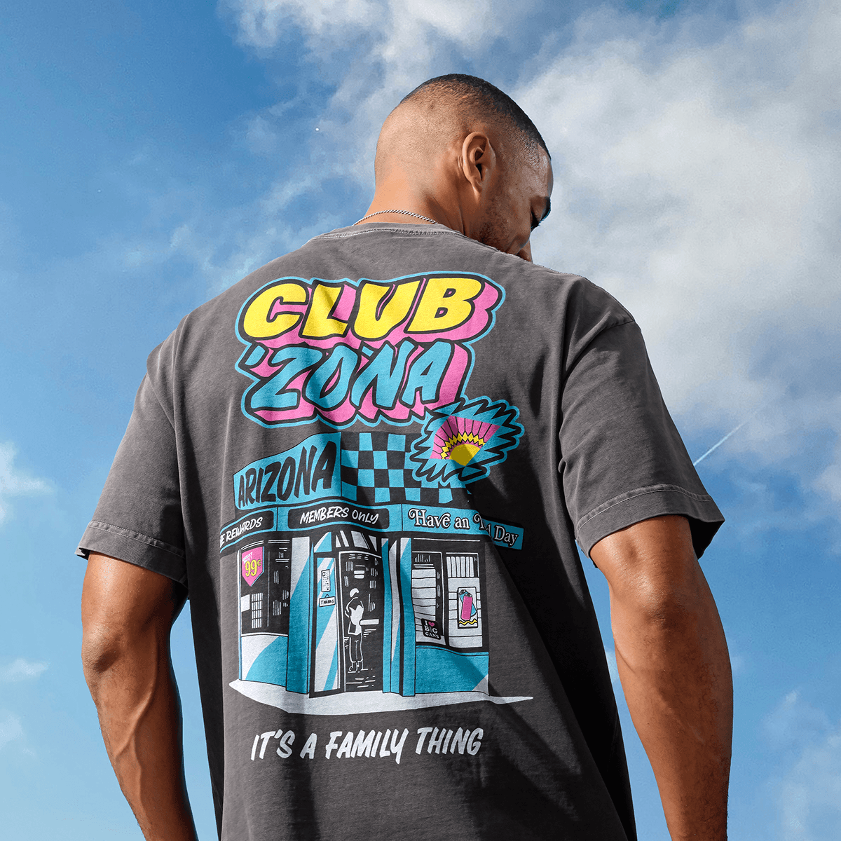 A man viewed from behind wearing a dark gray graphic t-shirt with a colorful CLUB ZONA design on the back, set against a blue sky with clouds.