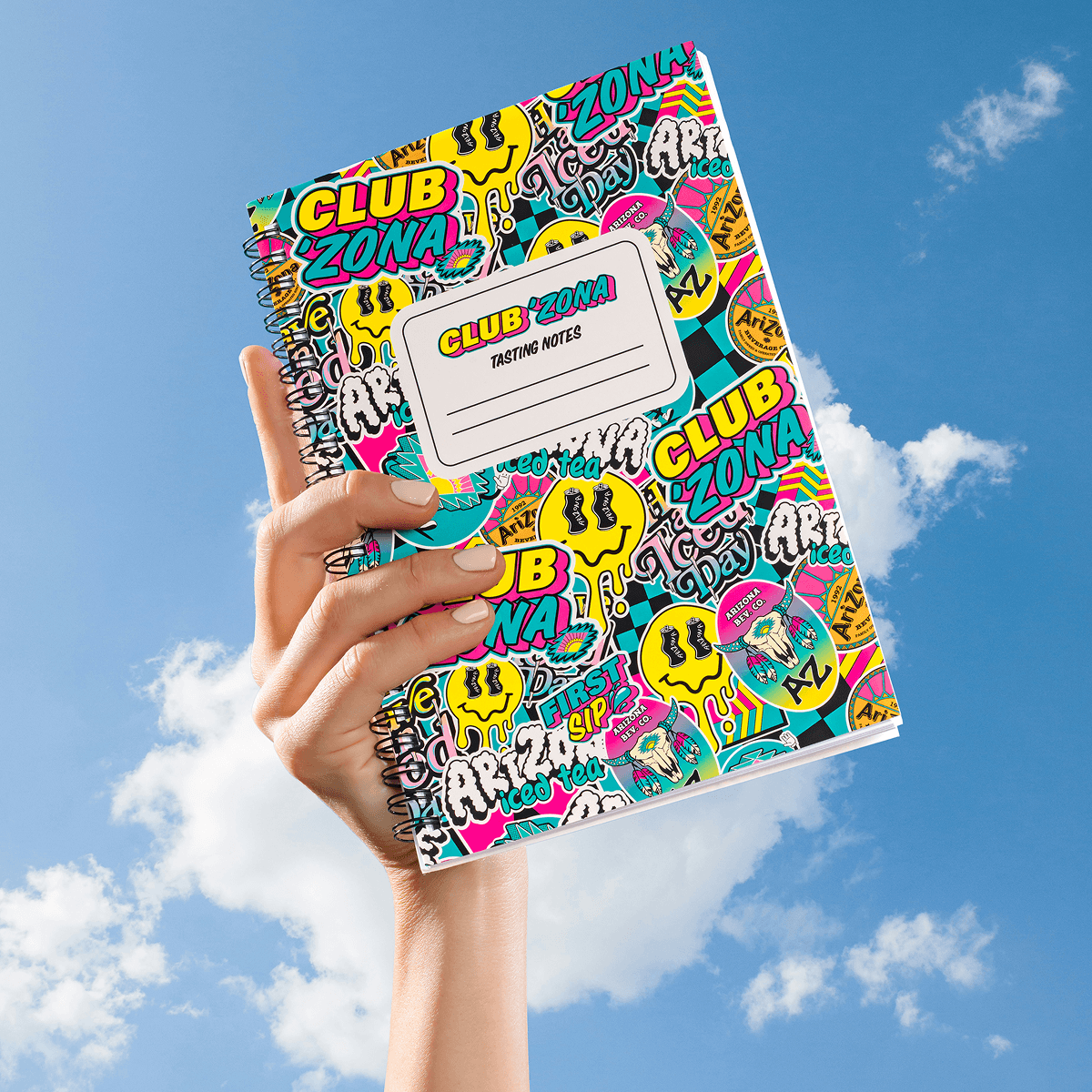A hand holding a spiral-bound notebook with a vibrant, multicolored graffiti-style cover that reads CLUB ZONA TASTING NOTES against a blue sky with clouds.