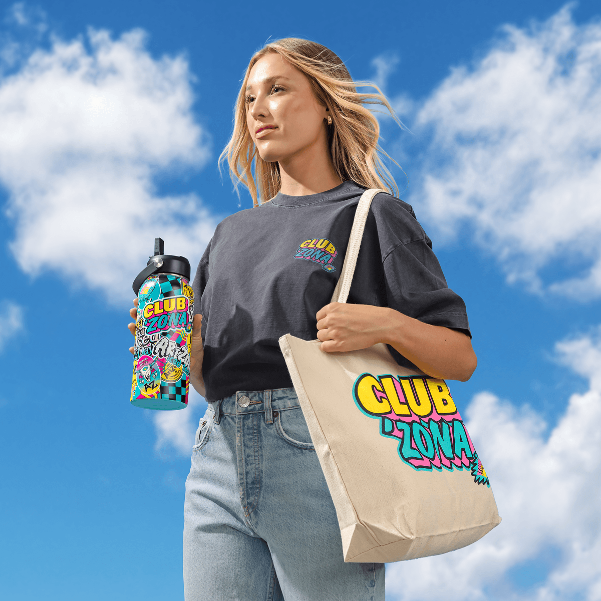A woman stands against a blue sky with clouds, holding a colorful water bottle and carrying a beige tote bag. Both the water bottle and tote bag feature a vibrant, multicolored retro-style graphic that reads CLUB ZONA