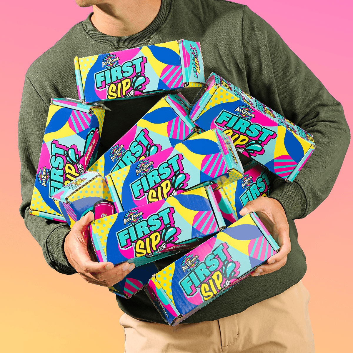 A person wearing an olive green sweatshirt and khaki pants holds a pile of colorful First Sip candy boxes against a pink-to-peach gradient background.