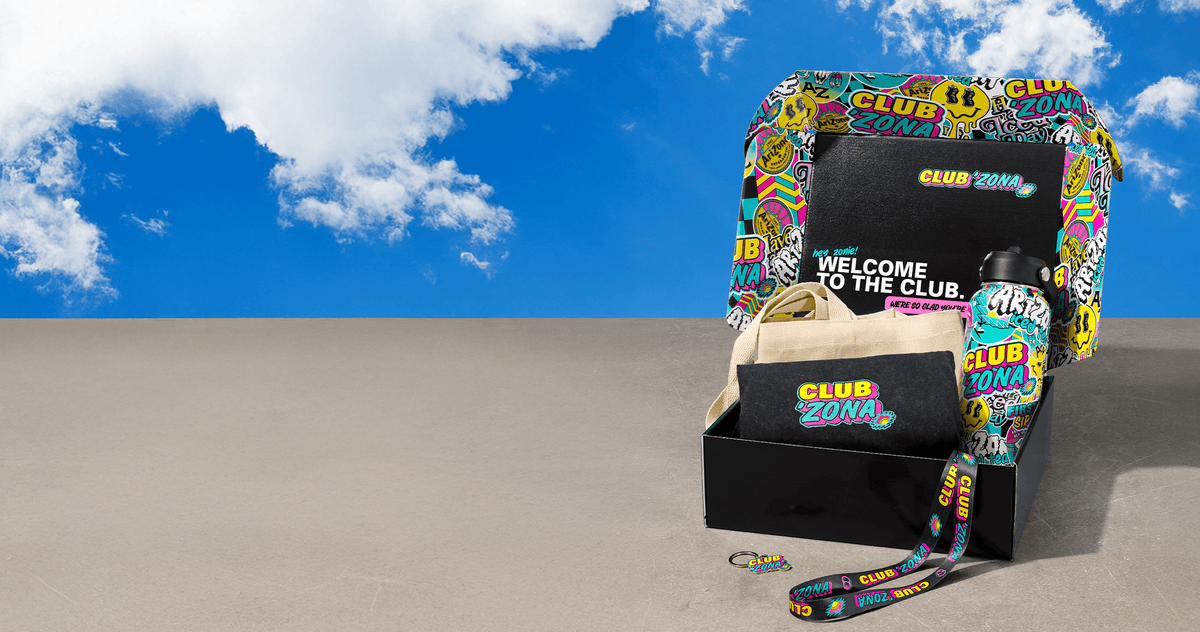 A Club Zona gift set displayed on a concrete surface under a bright blue sky with clouds. The set includes a black gift box with a colorful graffiti-patterned lid, a beige tote bag, a black t-shirt, a water bottle, and a wrist strap, all featuring the Club Zona logo.