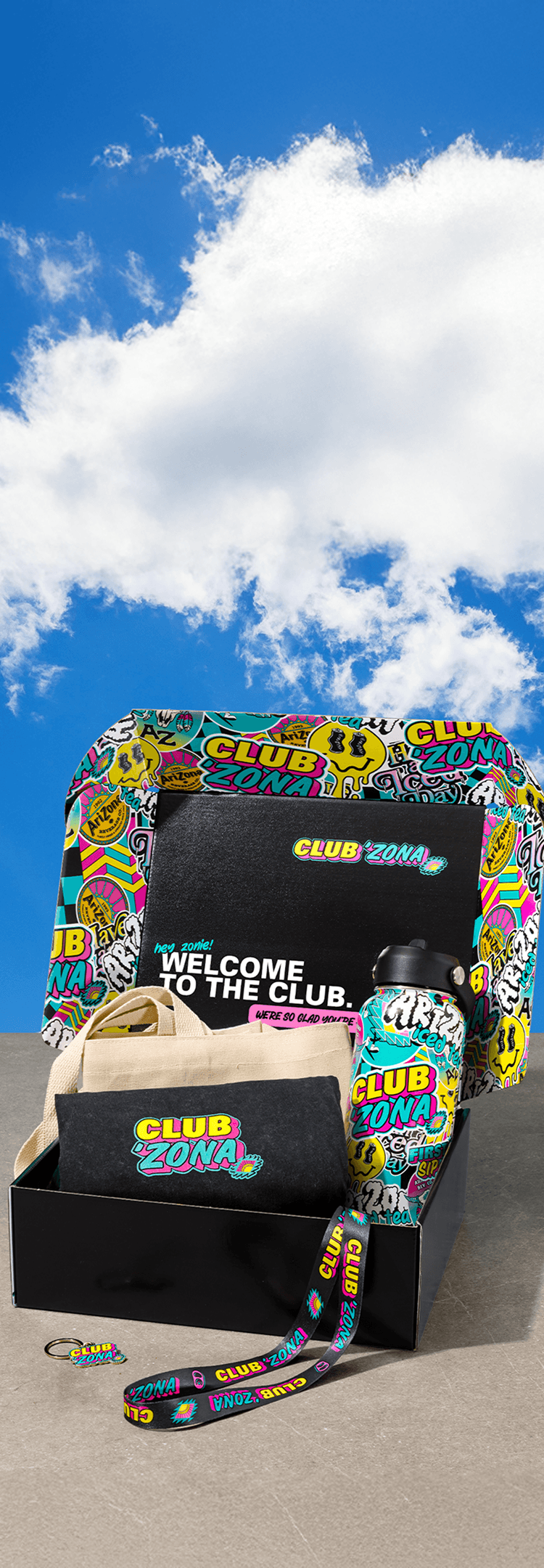 A Club Zona gift set displayed in an open black box on a sandy surface under a blue sky with clouds. The set includes a colorful graffiti-patterned tote bag, a black welcome pouch, a beige drawstring bag, a black bottle, and a matching wrist strap.