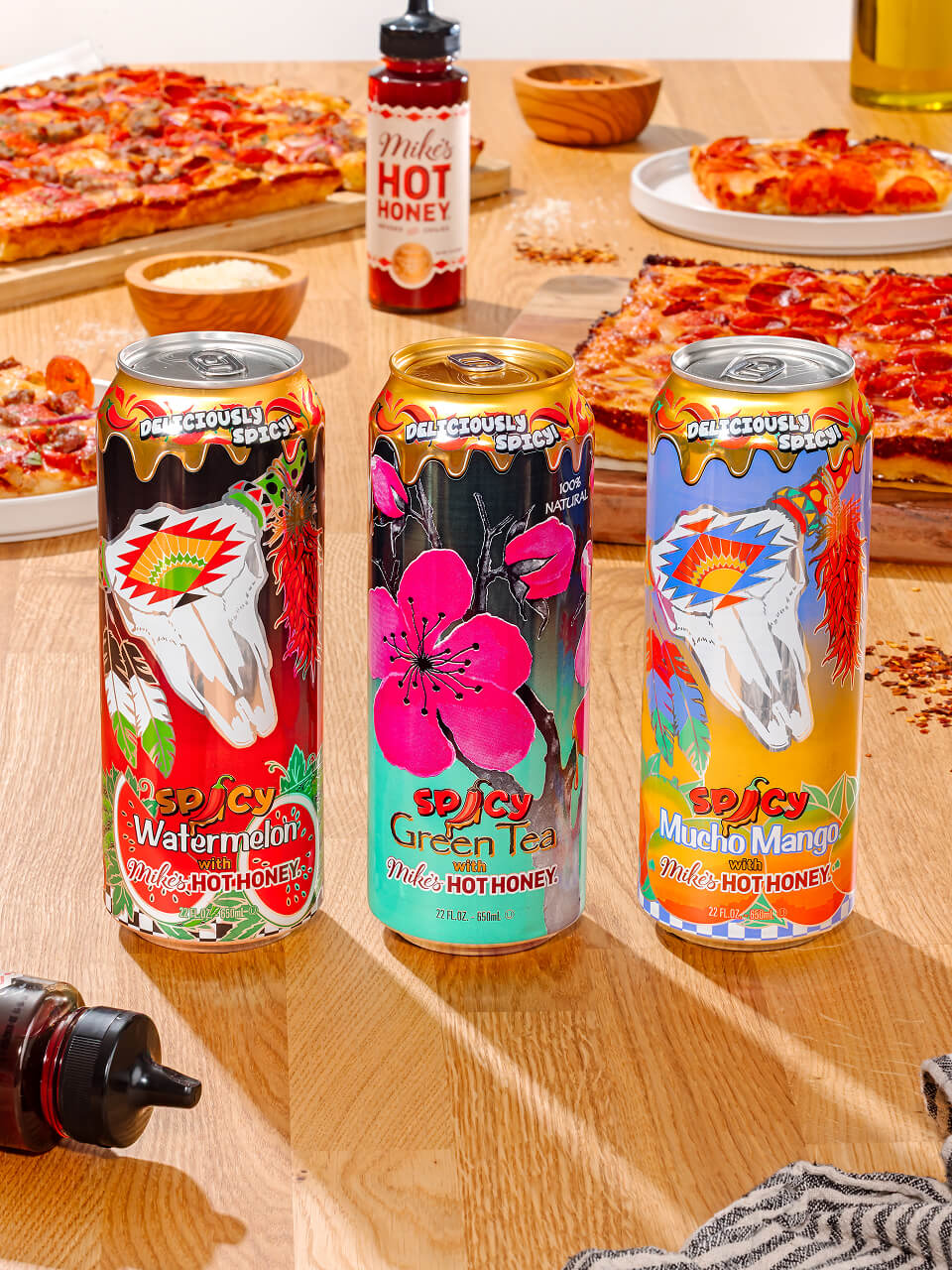Three colorful cans of Mike's Hot Honey on a wooden table with pizzas in the background