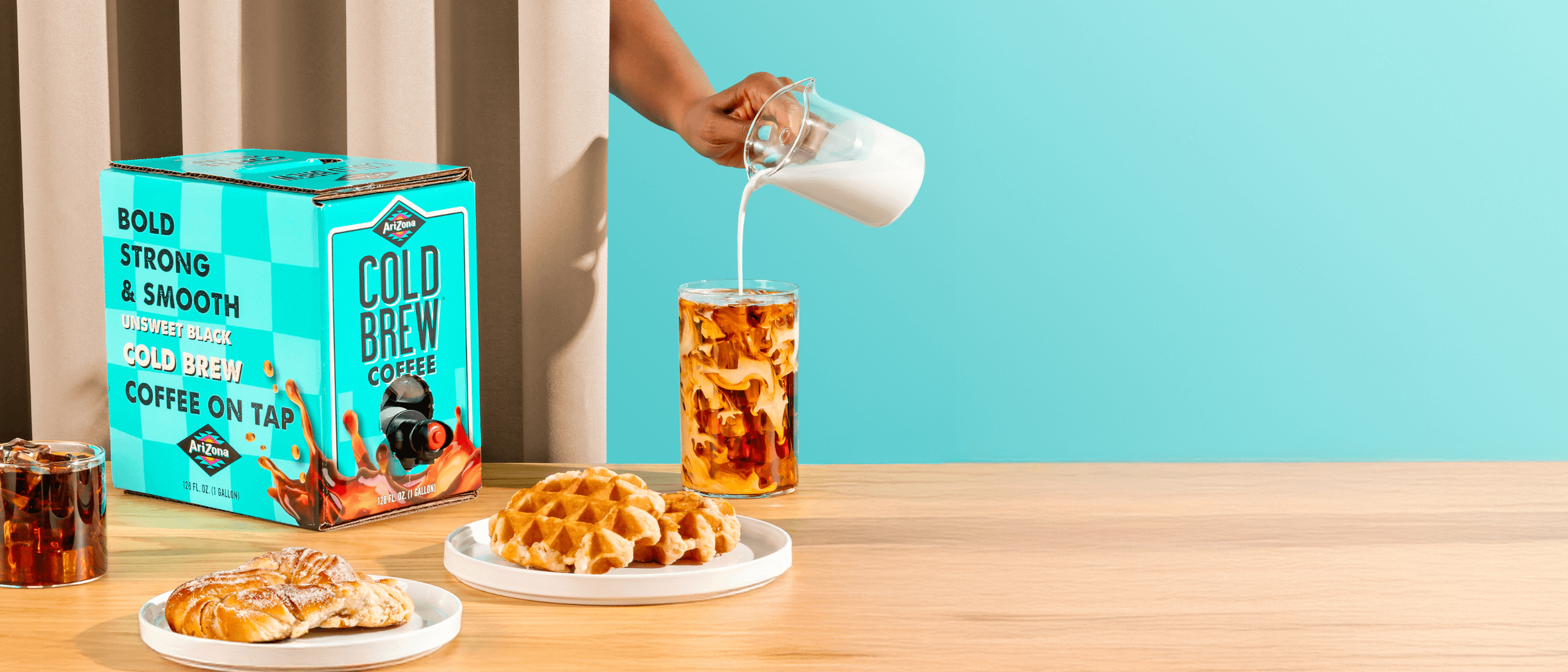 Person pouring milk into a glass of iced coffee next to a package of Folgers Cold Brew Coffee on a table with waffles.