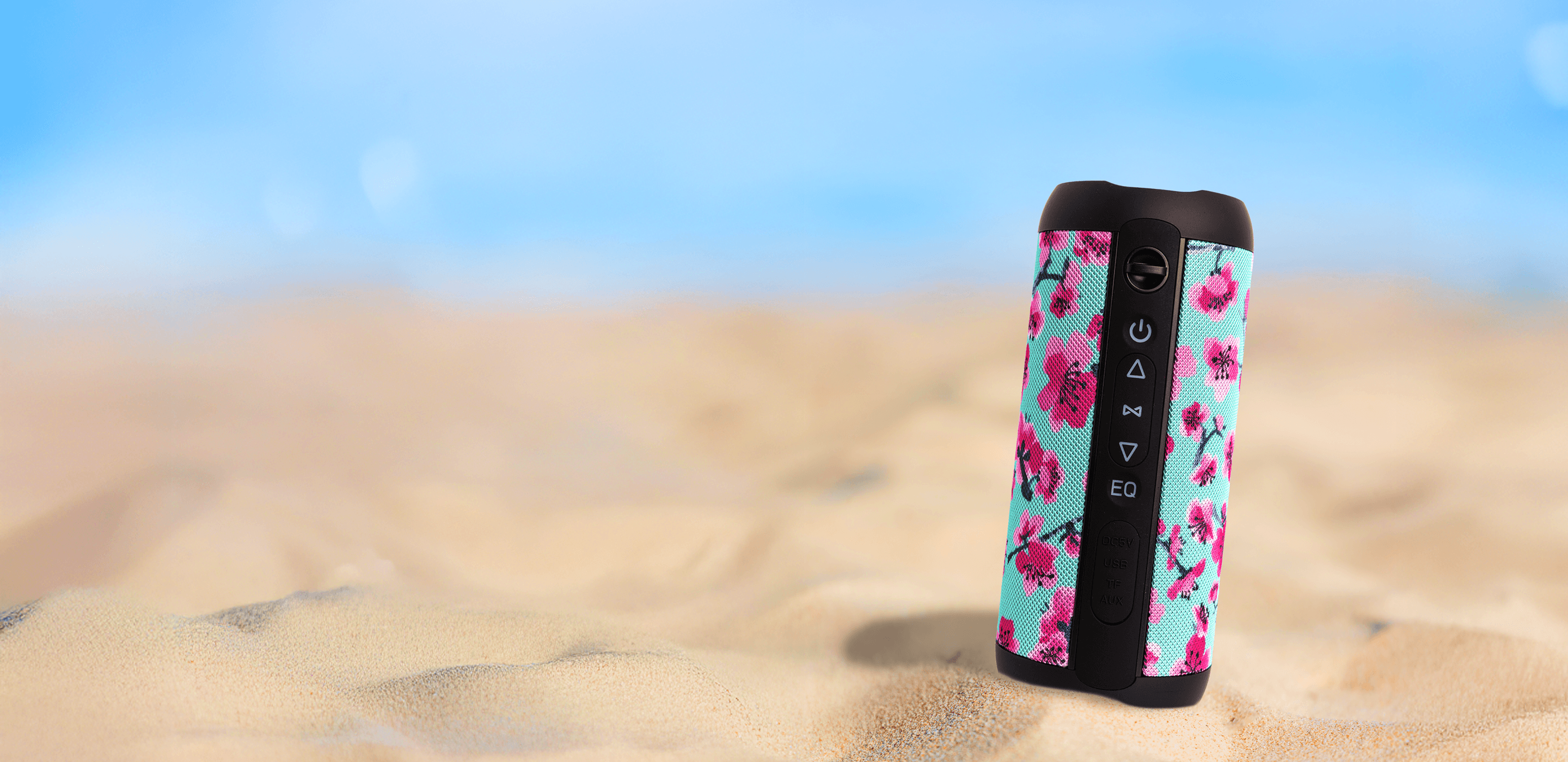A portable speaker with a  cherry blossom pattern on a light blue background, standing upright on a sandy beach with a blurred blue sky background.
