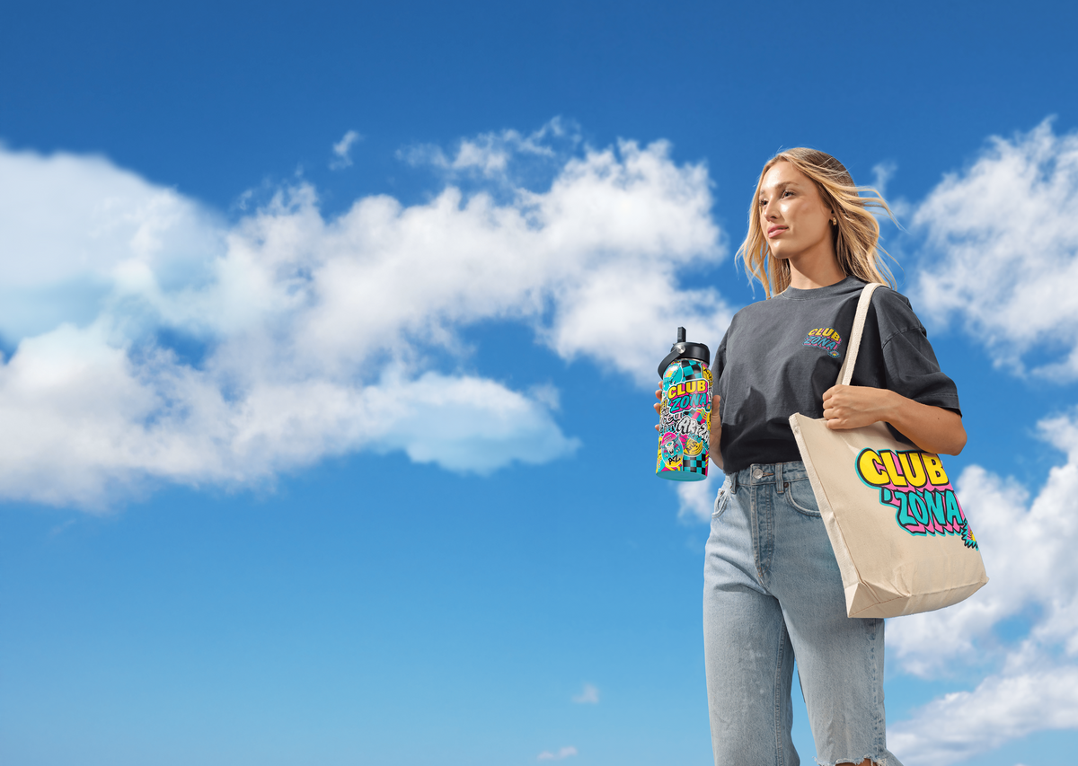 A woman stands against a bright blue sky with clouds, holding a colorful water bottle and carrying a beige tote bag printed with the words 'CLUB ZONA' in yellow, green, and blue. She wears a dark gray T-shirt and light blue jeans.