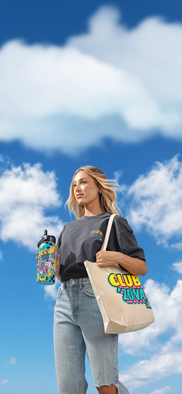 A woman stands outdoors against a blue sky with clouds, wearing a dark gray graphic T-shirt and light blue jeans. She carries a beige canvas tote bag with colorful 'CLUB ZONA' text and holds a colorful water bottle.